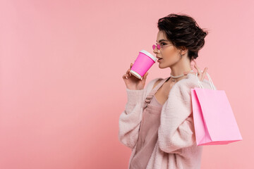 brunette woman in trendy sunglasses drinking coffee to go while holding shopping bag isolated on...