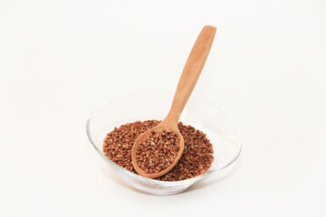 Small grains of natural brown buckwheat in a transparent glass plate and in a wooden spoon on a white background