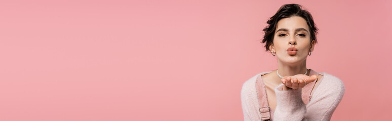 trendy brunette woman in warm cardigan looking at camera and sending air kiss isolated on pink,...