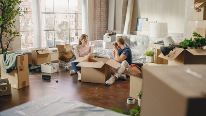 Obraz premium Happy Homeowners Moving In: Lovely Couple Sitting on the Floor of the Cozy Apartment Unpacking Cardboard Boxes, Taking Photos for Memories. Mortgage Loan, Real Estate. Nostalgia Cinematic Color