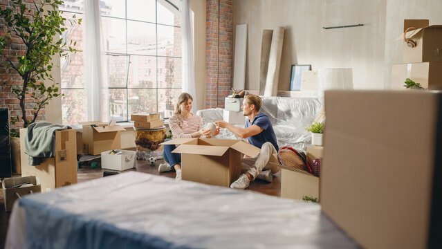 Happy Homeowners Moving In: Lovely Couple Sitting on the Floor of Cozy Apartment Unpacking Cardboard Boxes. Mortgage Loan, Real Estate, Sweet Home for Young Family. Bright Day full of Happy Memories