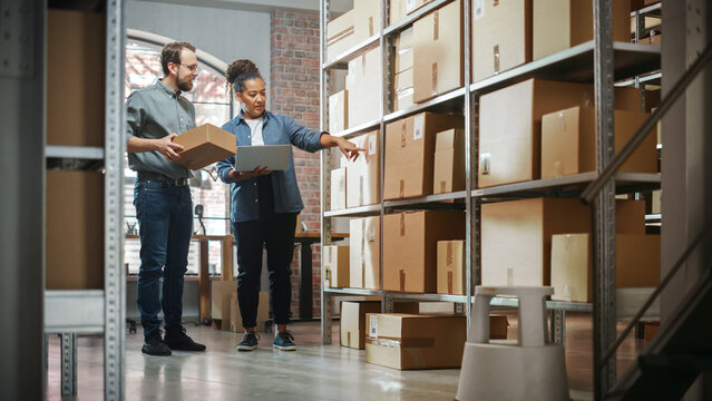 Small Business Owner And Employee Checking Stock And Inventory With Laptop Computer In Retail Warehouse Full Of Shelves With Goods. Working In Logistics, Online Shop Distribution Center.