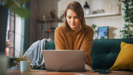 Fototapeta premium Portrait of Smiling Young Woman Working from Home on Laptop Computer in Sunny Cozy Apartment. Successful Creative Girl Entrepreneur Does Remote for e-Business Project, Online Shopping
