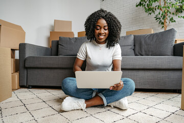 African woman with boxes in her new home sitting on the floor with laptop in the living room