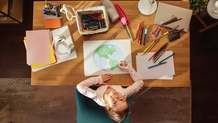 Fototapeta na wymiar Top View: Little Girl Drawing Our Beautiful Planet Earth, Looking up and Smiling. Talented Child Imagining Our Home Planet as a Happy Place with Clean, Sustainable Living. Cozy Sunny Day.