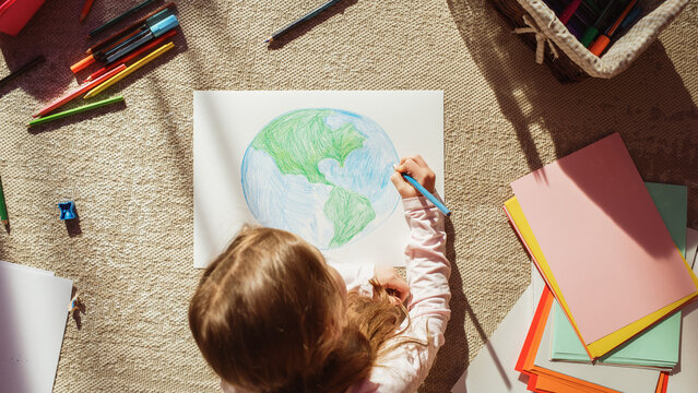 Top View: Little Girl Drawing Our Beautiful Planet Earth. Very Talented Child Having Fun at Home on the Floor, Imagining Our Home Planet as a Happy Place with Clean, Sustainable Living. Cozy Sunny Day