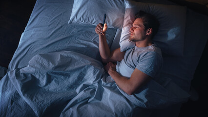 Top View Apartment Bedroom: Handsome Young Man Using Smartphone in Bed at Night. Smiling Guy Browsing Social Media, Dating Apps, Remote Work Software, Doing Internet Online Shopping on Mobile Phone