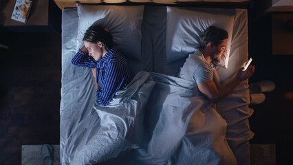 Top View Bedroom Apartment: Young Woman Uses Smartphone in Bed at Night When Her Male Partner Trying to Fall Asleep Beside. Couple Fight, Argue. Social Media, Doom Scrolling, Fake News Addiction