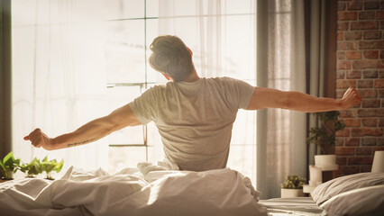 Handsome Young Man Waking up in the Morning, Stretches and Gets Out of Bed, Sun Shines From the Apartment Window in Bedroom, He is Ready for Business Opportunities, Achievements, Adventures - obrazy, fototapety, plakaty