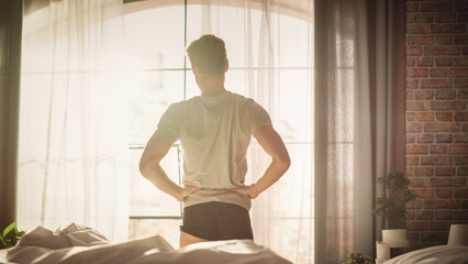 Fototapeta na wymiar Caucasian Young Man Waking up in the Morning, Stretches and Gets Out of Bed, Sun Shines From the Window, He is Ready for Business Opportunities, Achievements, Adventures. Seize the day