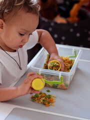 Little boy toddler playing with dry colorful pasta in tray while sitting at table in children room at home, selective focus, child learning and exploring sense of touch. Noodle sensory play for kids - obrazy, fototapety, plakaty