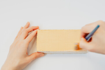 Hands painting a piece of wood with pale yellow acrylics on white background