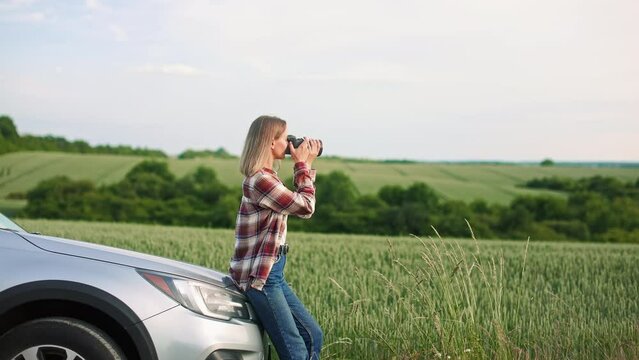 Side view of young, blonde lady sitting on car, taking photo by camera. Cheerful woman wearing plaid shirt and jeans, traveling by car, driving. Concept of art and photography.