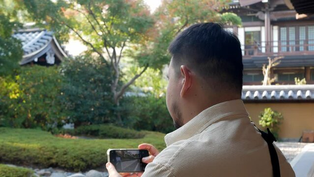asian man enjoy relaxing breathe and take photo with smartphone in japanese zen style garden in autumn daytime, relax peaceful
