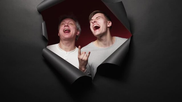 Hard rock joy. Funny men. Family entertainment. Expressive odd father and son showing horn gesture posing red background in black disrupted paper frame slow motion.