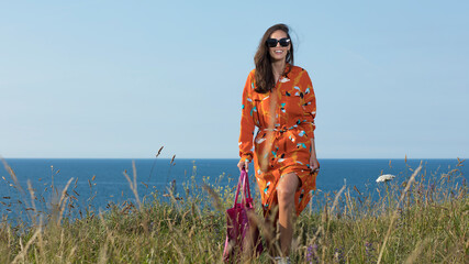 Spring outfits. Woman walking along the seaside outdoors smiling with a positive attitude and wearing sunglasses. Woman walking front in an orange dress bag overlooking the sea. Woman  after eating