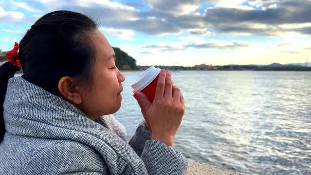A woman holding and drinking a cup of hot coffee by the sea during sunset and cold weather