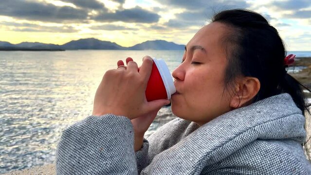A woman holding and drinking a cup of hot coffee by the sea during sunset and cold weather