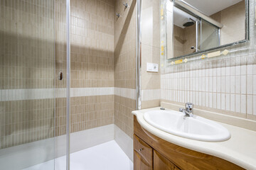 Bathroom with small white porcelain sink on white marble countertop and glass marble mirror with oak cabinets and glass-enclosed shower cubicle