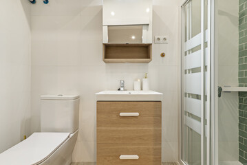 Fototapeta na wymiar Small bathroom with oak furniture with drawers, porcelain sink and shower cabin with glass partitions