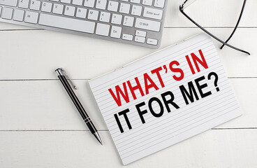 WHAT'S IN IT FOR ME text on notebook with keyboard , pen glasses on white wooden background
