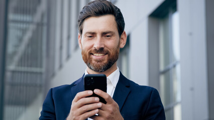 Smiling happy man holds phone browsing social network standing outdoors successful businessman...