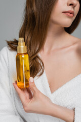 Fototapeta premium cropped view of young woman with shiny hair holding bottle with oil isolated on grey