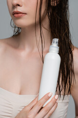 cropped view of young woman with bare shoulders holding spray bottle with treatment hair product isolated on grey