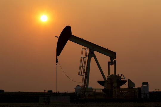 Silhouette of pumpjack with a glowing warm sun ball in the background, West of Airdrie; Alberta, Canada
