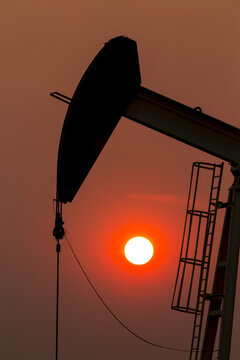 Silhouette of pumpjack with a glowing warm sun ball in the background, West of Airdrie; Alberta, Canada