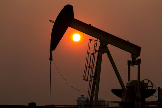 Silhouette of pumpjack with a glowing warm sun ball in the background, West of Airdrie; Alberta, Canada