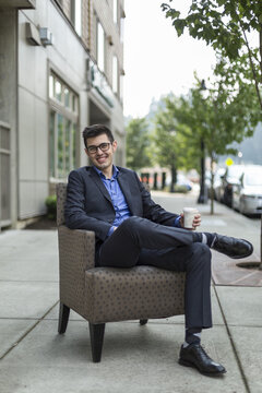 A young man sits in an armchair on an outdoor walkway and holds a cup of coffee; Bothell, Washington, United States of America