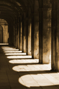 Walkway with shadows of arches and columns on the ground cast by bright sunlight; Venice, Italy