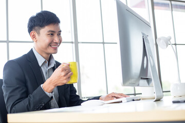 Asian business man drinking coffee sit at desk watching work project on computer. Male employee smiling looking at success work in the office
