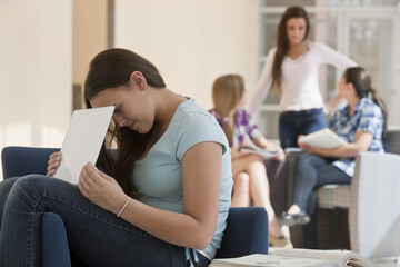 Teenage girl using laptop at school and hiding the screen from others with two students looking at textbooks in the background