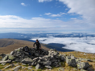 Man standing in wind protection construction made of rocks near the summit cross of Zirbitzkogel, Seetal Alps in Murtal, Styria, Austria, Europe. Panoramic view on the surrounding mountain ranges