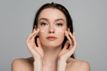 young woman with wet hair applying face cream on cheeks isolated on grey