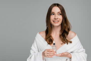 happy young woman with shiny hair holding bottle with serum isolated on grey