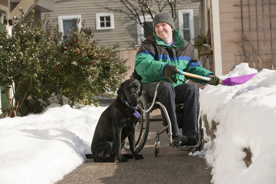 Woman With Multiple Sclerosis Shoveling Snow In A Wheelchair With A Service Dog