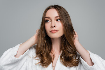 young woman in bathrobe touching shiny and healthy hair while looking away isolated on grey - obrazy, fototapety, plakaty