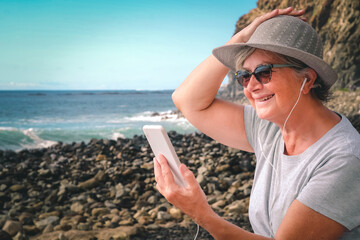 Cheerful senior woman sitting at the beach holding his hat while listening music by smartphone -...