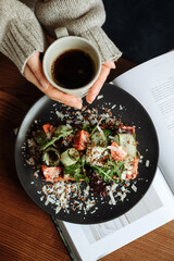 Females hand holds a cup of black coffee near the plate with salad