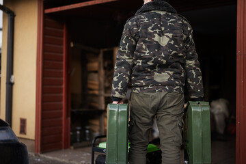 Back of man wear military jacket hold two canisters with gasoline against portable mobile backup...