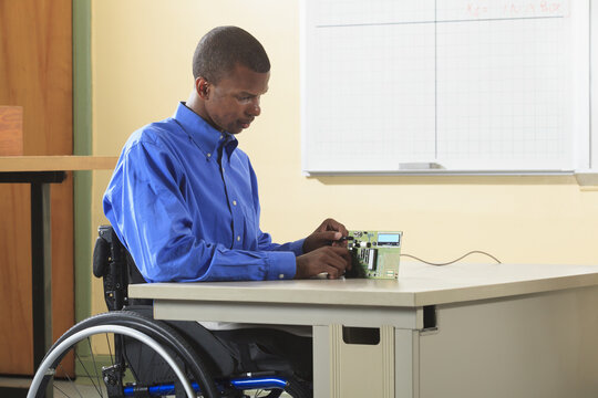 Engineering student sitting in a wheelchair examining microprocessor circuit board for electronics lab experiment