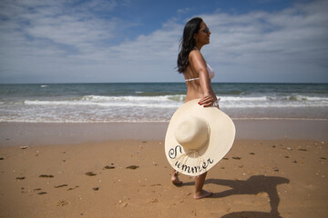 Fototapeta premium South American woman, young and beautiful, brunette with bikini and hat with the word summer held in her hand, on her back, running. Concept sea, sand, sun, beach, vacation, travel. Selective focus.