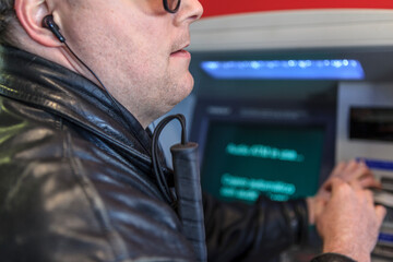 Man with visual impairment standing at a bank machine with a white cane and earphones