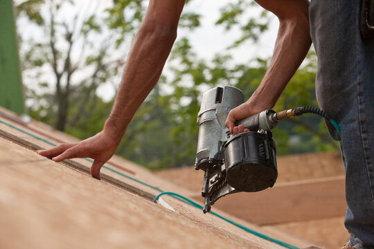 Hispanic carpenter using a nail gun on the roof panel of a house under construction