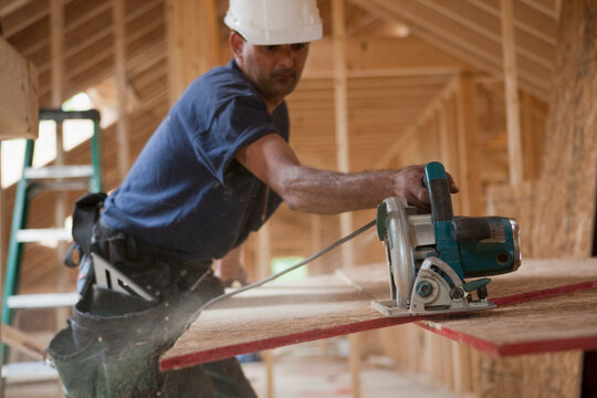 Hispanic carpenter trimming roof panel with a circular saw at a house under construction