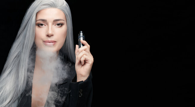Sensual gray haired woman using electronic cigarette. Beauty model woman with a vaping device blowing a white cloud of smoke. Horizontal portrait isolated on black background with copy space