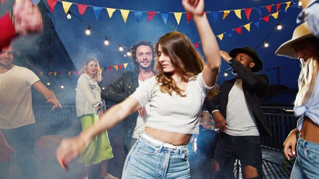 Group of happy young relaxed people having party on rooftop. Beautiful man and woman dancing in centre of circle with positive cheerful friends listening to music having fun at weekends.